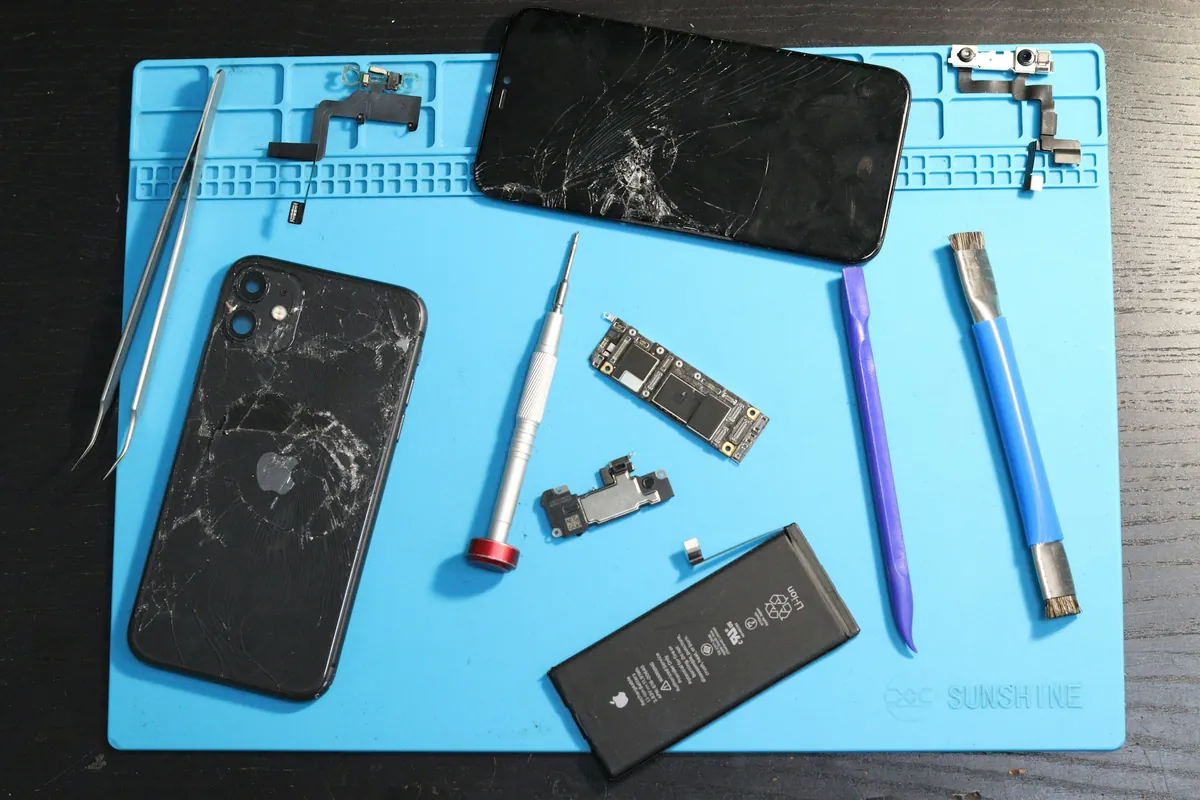 iPhone à l'écran fissuré et batterie posés sur un tapis d'atelier avec des tournevis